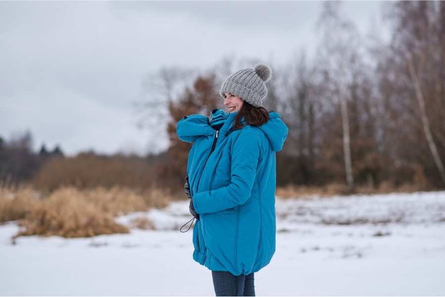 Smiling mom with a beanie on her head standing by a snowy plain with her baby tucked under a warm turquoise baby carrier